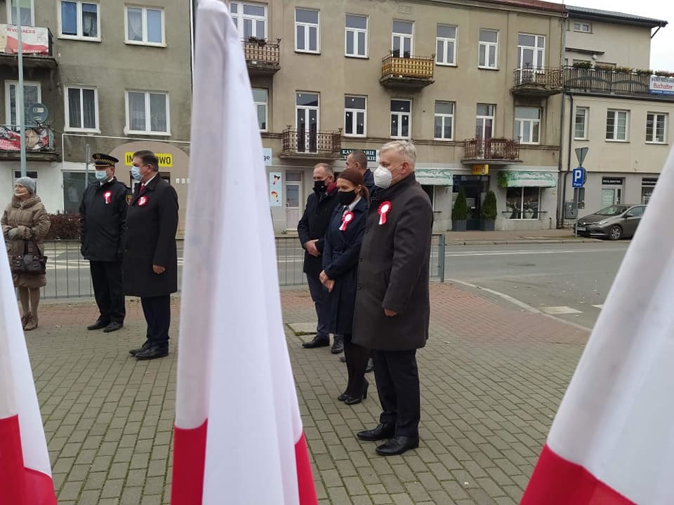 Przedstawiciele powiatu płońskiego i urzędu miasta stoją na chodniku na tle kamienic na pierwszym planie flagi Polski.jpg
