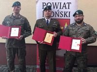 Przedstawiciele jednostek strzeleckich stoją na tle rollupu powiatu płońskiego i trzymają pamiątkowe tabliczki gratulacyjne