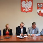 Na zdjęciu pierwszy od lewej siedzi mężczyzna, druga od lewej siedzi kobieta, pośrodku siedzi mężczyzna, pierwszy od prawej siedzi mężczyzna, drugi od prawej siedzi mężczyzna, w tle godło Polski i herb Powiatu.