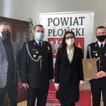 Starosta i Wicestarosta stoją z przedstawicielami OSP Nowe Miasto na tle rollupu powiatu płońskiego.jpg
