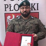 Przedstawiciel jednostki strzeleckiej stoi na tle rollupu powiatu płońskiego i trzyma pamiątkową tabliczkę gratulacyjną
