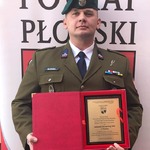 Przedstawiciel jednostki strzeleckiej stoi na tle rollupu powiatu płońskiego i trzyma pamiątkową tabliczkę gratulacyjną